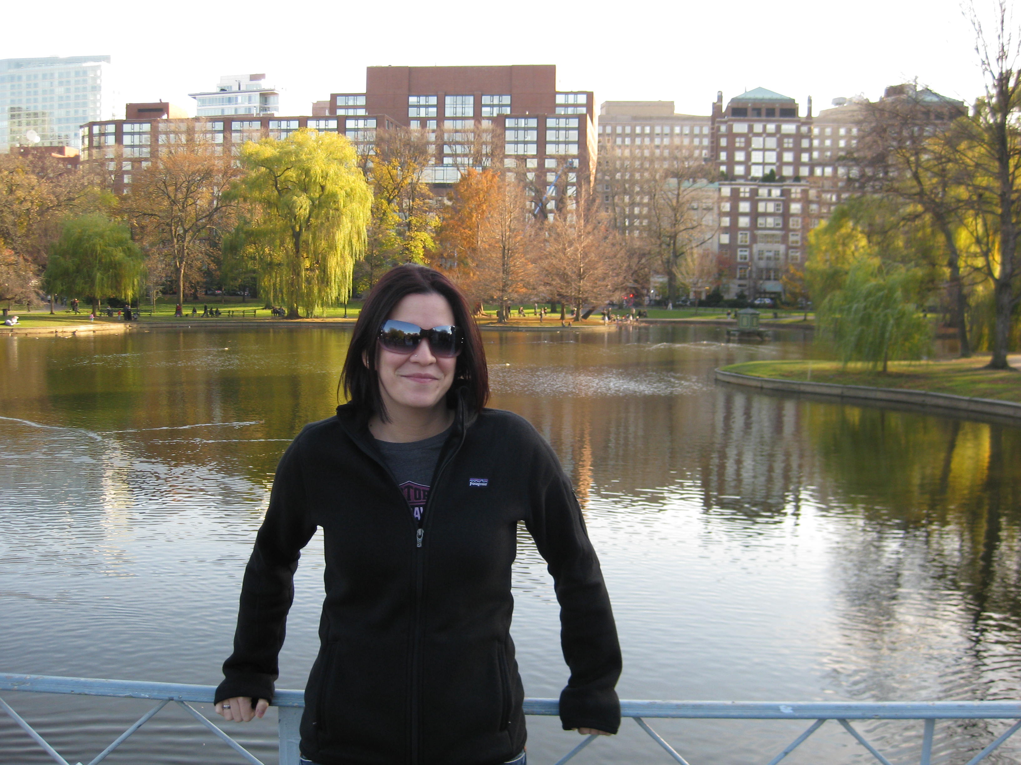 A person with short brunette hair wearing a black fleece sweatshirt, wearing sunglasses and standing in front of a body of water with green and orange trees in the background.