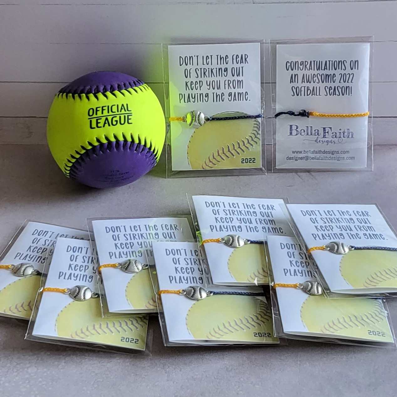 A purple and yellow softball next to team favors of cute string bracelets with softball charms.