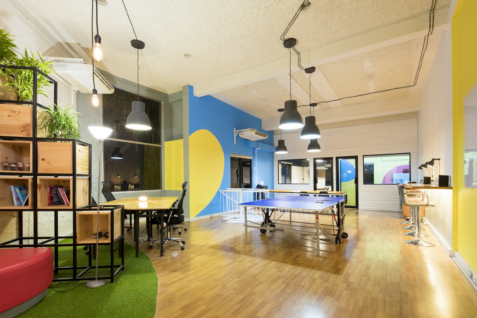 A colorful work office with a bright blue and yellow accent wall, wooden floors, and black and gray hanging ceiling lights.